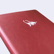 Close up of debossed detailing of 2 dinosaurs on the cover of a burgundy leather sketchbook.