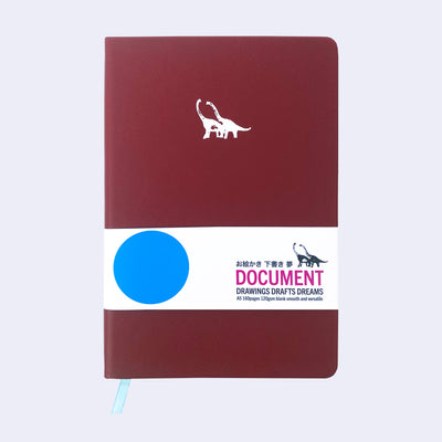 Burgundy pleather notebook with a small embossment of a pair of long necked dinosaurs, in the upper center of journal. Paper sleeve reads "Document Drawings Drafts Dreams."