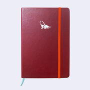 Burgundy pleather notebook with a small embossment of a pair of long necked dinosaurs, in the upper center of journal. A bright orange elastic band holds the notebook closed.