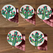 5 circular ceramic brooches, collaged together into one image. Each brooch has a cream colored base and carved illustration of a green cactus with red blossoms.