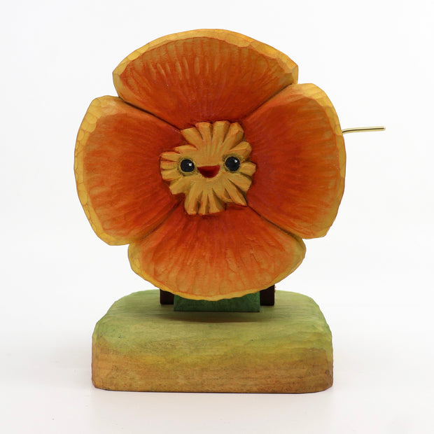 Small wooden sculpture of a orange poppy with a small cartoonish face. Sculpture is on a stand and has a lever attached to the back.