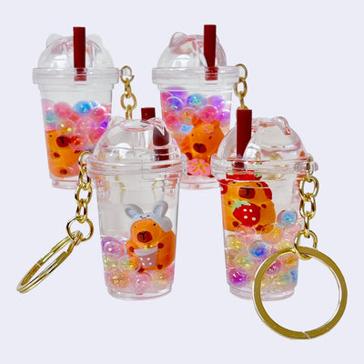 4 clear to-go cup shaped containers with colorful balls inside and a capybara figure, varying in design with floral or animal themes. Each cup has a plastic straw attached and are attached to a gold hardware keyring.