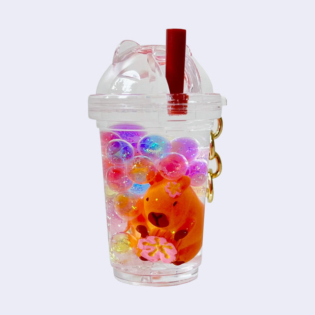 Keychain of a clear to-go cup with colorful glitter filled balls inside and a capybara figure, holding a pink flower. Charm is attached to a gold hardware keychain.