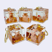 5 different clear cubes with gold glitter leaves inside and small figures of closed eye capybaras, sitting or laying in the center. Each cube is attached to a gold hardware keyring.