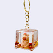 Clear cube with gold glitter leaves inside and small figure of closed eye capybara, sitting upright with a tangerine atop its head. Cube is attached to a gold hardware key chain.