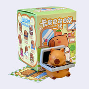 Blind box figure of a capybara, cute and chubby in a cartoon style sitting on the ground with its head half through a laptop. It sits in front of its product packaging.