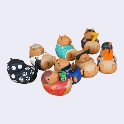 8 differently dressed figures of capybaras, laying on their backs with their heads slightly curled towards their stomachs. Some hold props such as the number 7, a ukelele, alcohol, etc.