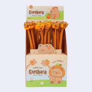 3 gel pens with brown rubber bodies and pen toppers of cute, cartoon capybaras each in a different pose. One sits atop an orange, another clings to the side of an orange and the final sits with an orange hood hat on. Pens are in their cardboard display.