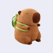 Plush doll of a brown capybara with dark brown ears, paws and snout and a cute closed eye expression. It wears a green backpack shaped like a turtle, which is removable. 