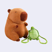 Plush doll of a brown capybara with dark brown ears, paws and snout and a cute closed eye expression. It sits next to a green turtle shaped backpack, which can be worn or removed.