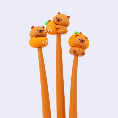 3 gel pens with brown rubber bodies and pen toppers of cute, cartoon capybaras each in a different pose. One sits atop an orange, another clings to the side of an orange and the final sits with an orange hood hat on. 