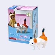 Small plastic figure of a calico cat with a blocky body, as if carved out of wood then painted. Behind is its product packaging.