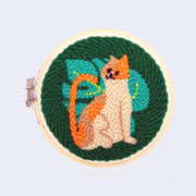 Final product for a Punch Needle Kit with the design of an orange cat with a large Monstera style leaf behind it on a dark green background.