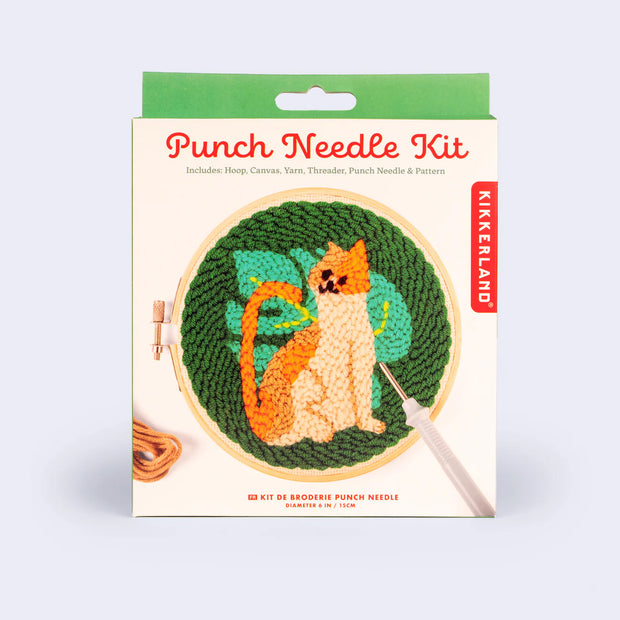 Product packaging for a Punch Needle Kit with the design of an orange cat with a large Monstera style leaf behind it on a dark green background.