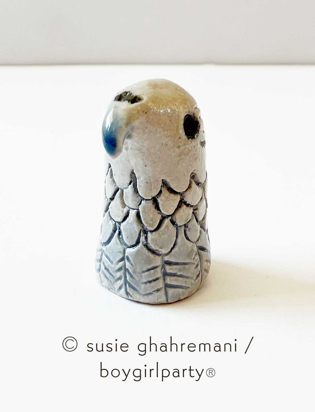 Small ceramic figure of a eagle head with a rounded wing pattern and a curved beak and deep set eye.