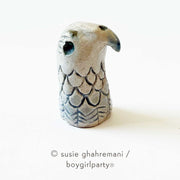 Small ceramic figure of a eagle head with a rounded wing pattern and a curved beak and deep set eye.