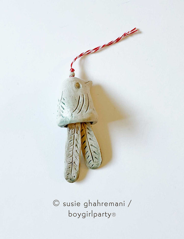 Off white ceramic bell with the top made to look like a bird and the bell part 2 feathers. Attached to the top is a red and white twine.