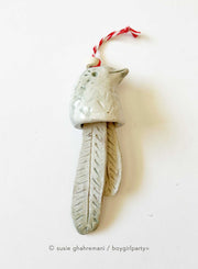 Off white ceramic bell with the top made to look like a bird and the bell part 2 feathers. Attached to the top is a red and white twine.