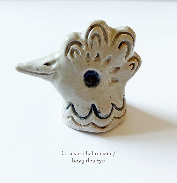 Off white ceramic sculpture of a chicken head, with a smiling beak, ruffled head feathers and an in set eye.