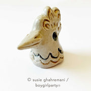 Off white ceramic sculpture of a chicken head, with a smiling beak, ruffled head feathers and an in set eye.