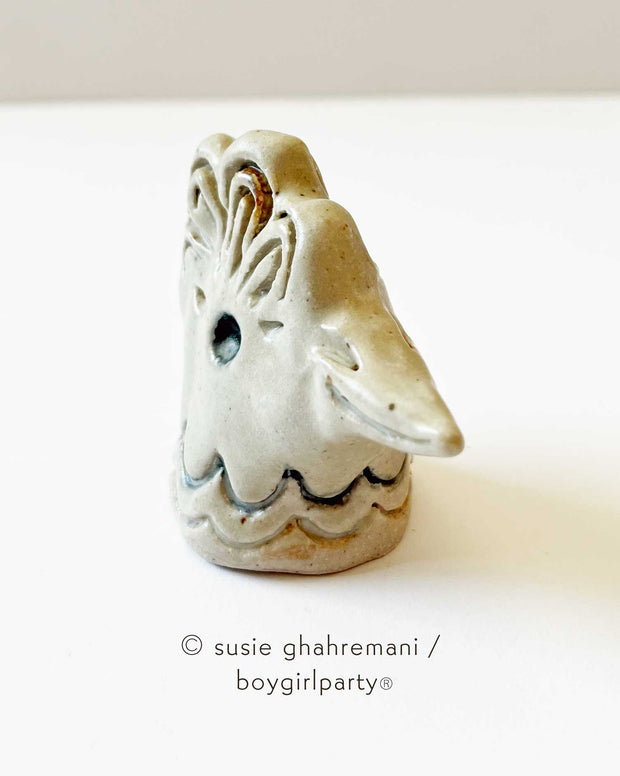 Off white ceramic sculpture of a chicken head, with a smiling beak, ruffled head feathers and an in set eye.