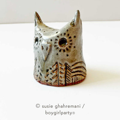 Brown toned ceramic sculpture of an owl head with pointed ears, a flat beak and patterned trees and dots on its body.