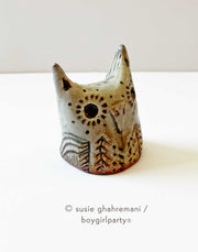 Brown toned ceramic sculpture of an owl head with pointed ears, a flat beak and patterned trees and dots on its body.