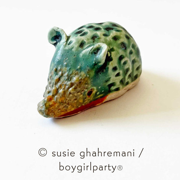 Small ceramic sculpture of a bluish green hedgehog with brown snout and brown bottom. It has a textured body, small pointed snout and small round ears.