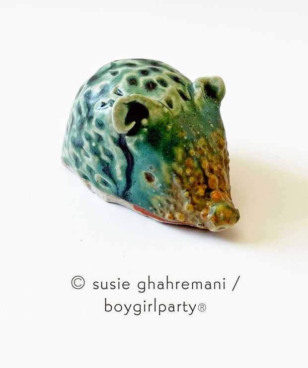 Small ceramic sculpture of a bluish green hedgehog with brown snout and brown bottom. It has a textured body, small pointed snout and small round ears.