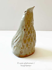 Ceramic sculpture of a bird, with a pointed cone shaped body. It has textured feathers and a pointed beak that looks upward.