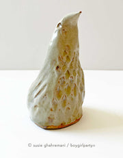 Ceramic sculpture of a bird, with a pointed cone shaped body. It has textured feathers and a pointed beak that looks upward.