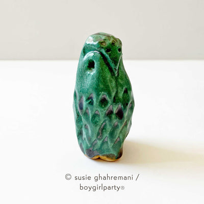 Small ceramic sculpture of a turquoise colored owl, standing with simplistic features and carved out feathers.