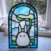 Stained glass small flat arch shaped piece, with blue glass around the border and a cute, small white bunny like character in the middle. It holds a leaf umbrella over its head.