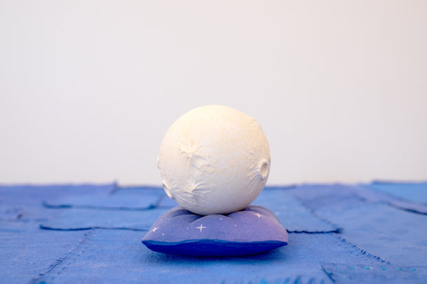 Small carved wooden sculpture of a white moon with a closed eye, calm smiling expression. It rests atop of a purple wooden pillow with star patterns, carved to look soft.