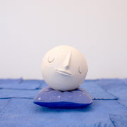 Small carved wooden sculpture of a white moon with a closed eye, calm smiling expression. It rests atop of a purple wooden pillow with star patterns, carved to look soft.