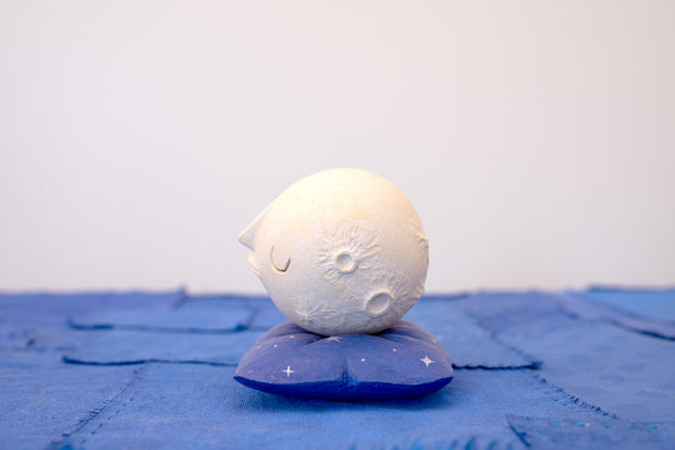 Small carved wooden sculpture of a white moon with a closed eye, calm smiling expression. It rests atop of a purple wooden pillow with star patterns, carved to look soft.