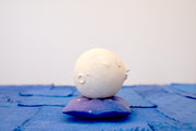 Small carved wooden sculpture of a white moon with a closed eye, calm smiling expression. It rests atop of a purple wooden pillow with star patterns, carved to look soft.