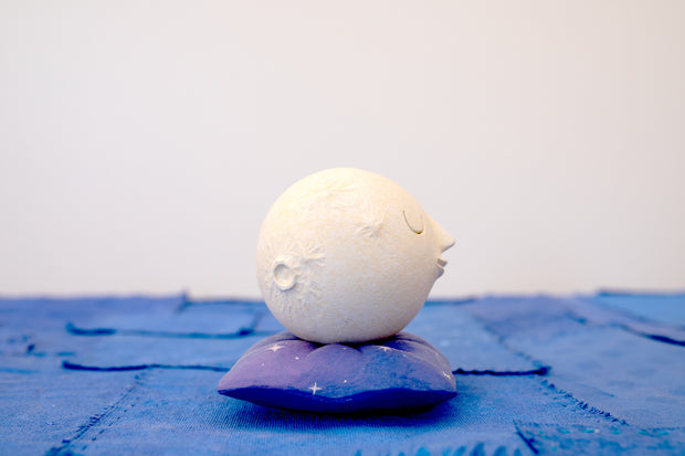 Small carved wooden sculpture of a white moon with a closed eye, calm smiling expression. It rests atop of a purple wooden pillow with star patterns, carved to look soft.