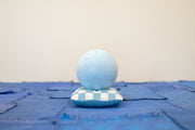 Small carved wooden sculpture of a blue moon with a closed eye, calm smiling expression. It rests atop of a white and blue grid patterned wooden pillow, carved to look soft.