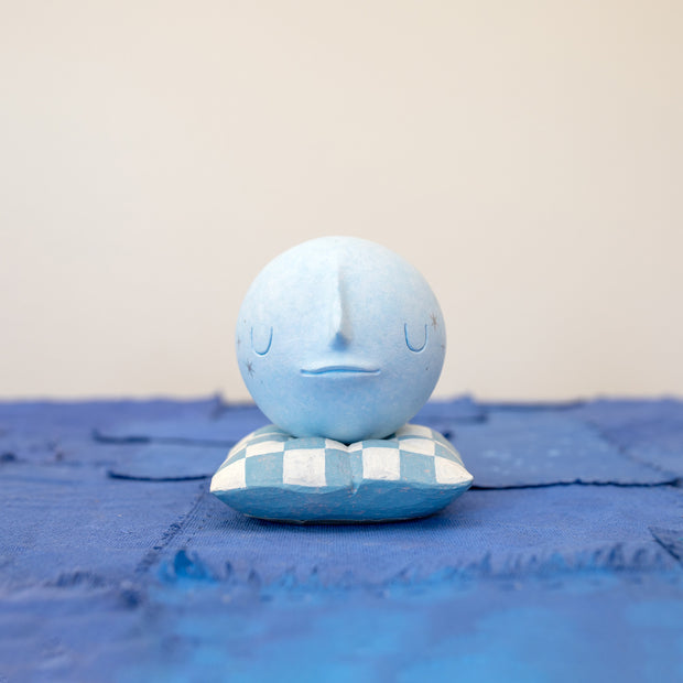 Small carved wooden sculpture of a blue moon with a closed eye, calm smiling expression. It rests atop of a white and blue grid patterned wooden pillow, carved to look soft.