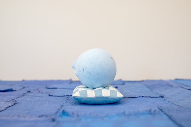 Small carved wooden sculpture of a blue moon with a closed eye, calm smiling expression. It rests atop of a white and blue grid patterned wooden pillow, carved to look soft.