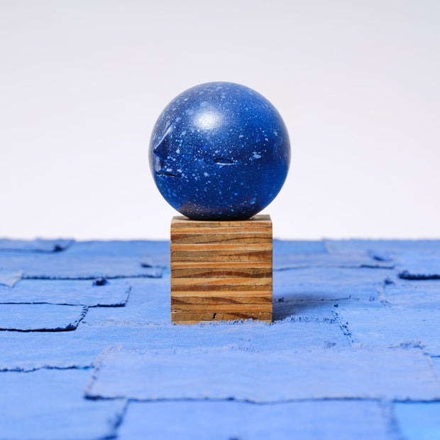 Small carved wooden sculpture of a dark blue moon with a closed eye, calm smiling expression. It rests atop of a striated grain wooden square pedestal. 