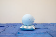 Small carved wooden sculpture of a blue moon with a closed eye, calm smiling expression. It rests atop of a white and blue grid patterned wooden pillow, carved to look soft.