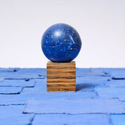 Small carved wooden sculpture of a dark blue moon with a closed eye, calm smiling expression. It rests atop of a striated grain wooden square pedestal. 