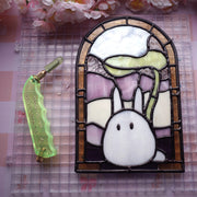 Stained glass artwork of a small white ChibiTotoro, holding a leaf above its head and encased in an arch shaped glass framing with shades of green, orange, pink and purple.