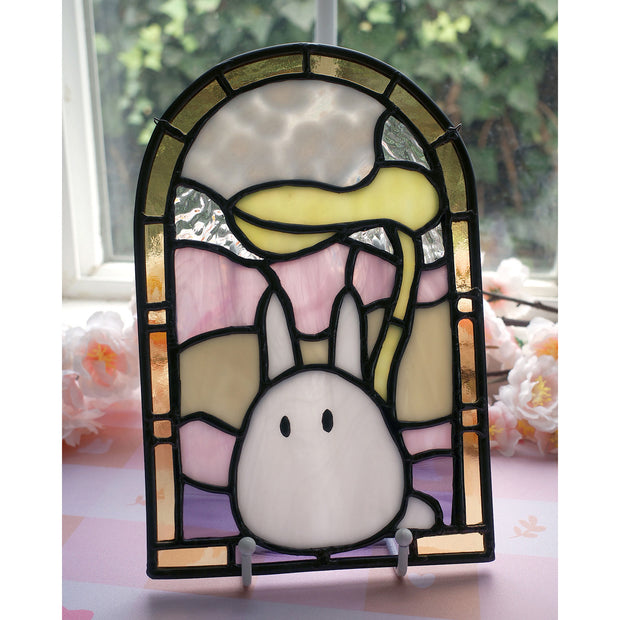 Stained glass artwork of a small white ChibiTotoro, holding a leaf above its head and encased in an arch shaped glass framing with shades of green, orange, pink and purple.