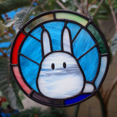 Circular stained glass hanging with a rainbow colored exterior circle, and interior blue background and a mostly white Chibi Totoro character.