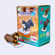 Figure of a small brown dachshund leaning forward on its front paws, pushing forward a plush cleaning pad. It stands next to its product packaging.