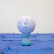 Small carved wooden sculpture of a purple moon with a closed eye, calm smiling expression. It rests atop of a blue wooden vessel shaped pedestal, like a kokeshi doll.