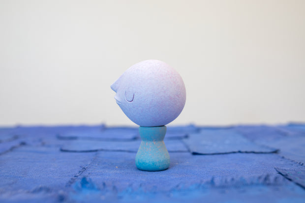 Small carved wooden sculpture of a purple moon with a closed eye, calm smiling expression. It rests atop of a blue wooden vessel shaped pedestal, like a kokeshi doll.